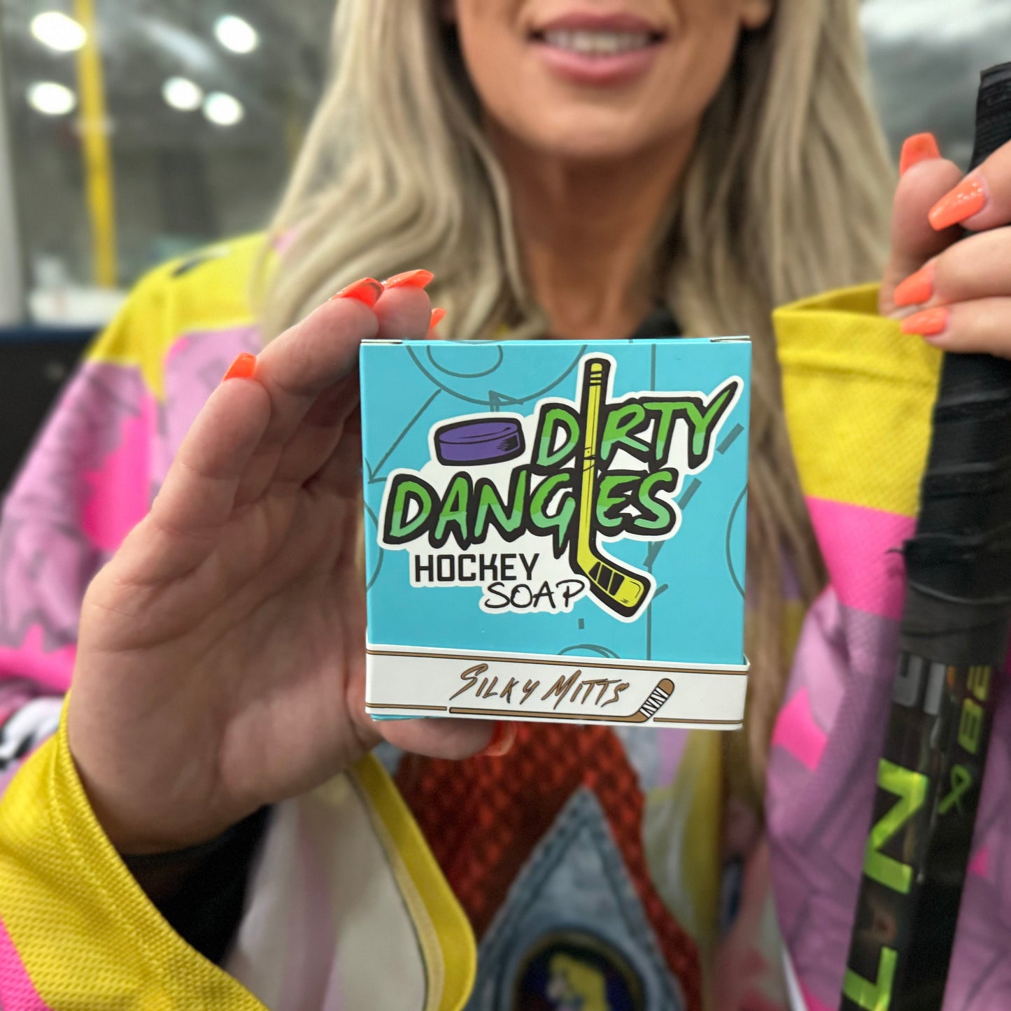 a woman hockey player holding a blue box of dirty dangles hockey soap silky mitts scent inside an ice rink.