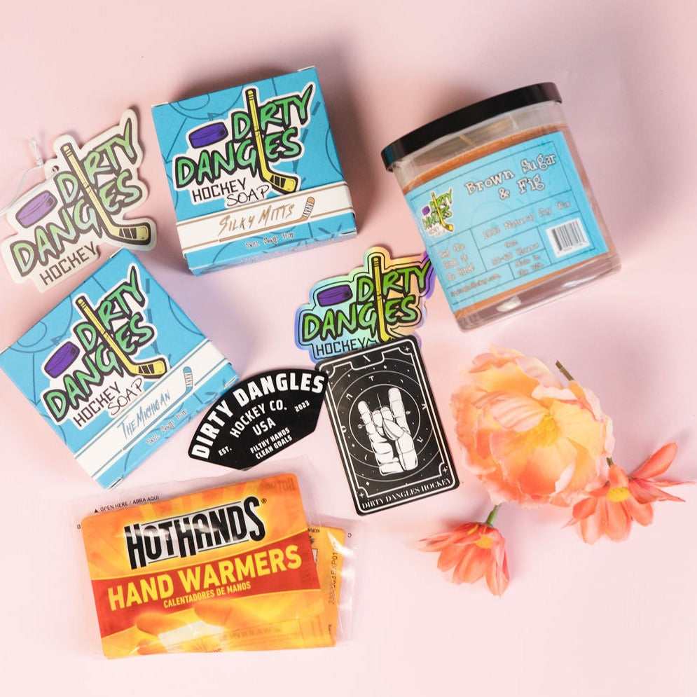A dirty dangles hockey mom bundle on a pink background with flowers. 2 natural hocky soap bars, a scented candle, car air freshener, hand warmers and stickers
