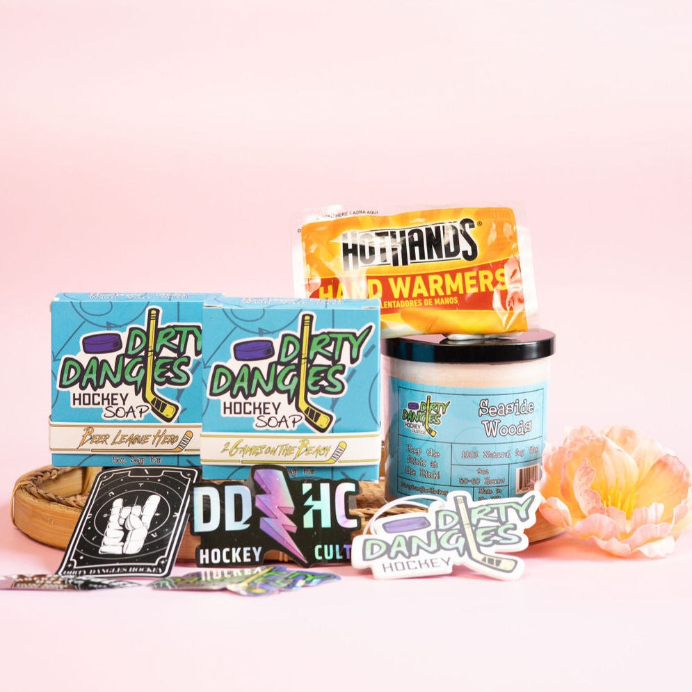A dirty dangles hockey mom bundle on a pink background with flowers. 2 natural hocky soap bars, a scented candle, car air freshener, hand warmers and stickers