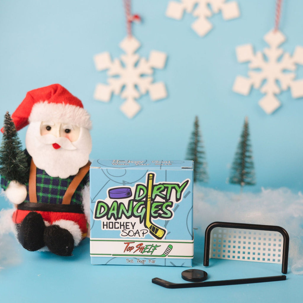 A bar of dirty dangles Top Shelf soap on a blue Christmas background with a stuffed Santa Claus and a tiny hockey goal, puck and stick.