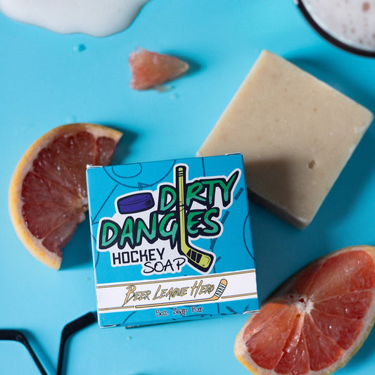 A bar of dirty dangles hockey soap on a blue background with a pale ale, grapefruit and some toy hockey sticks. Dirty dangles hockey soap