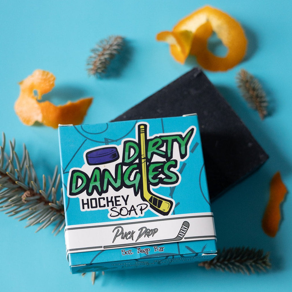 A bar of dirty dangles hockey soap puck drop scent on a blue background with pine and orange peel.