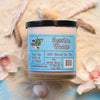 A beige dirty dangles hockey candle seaside woods scent on a blue background with sand, seashells and flowers. Seaside Woods.