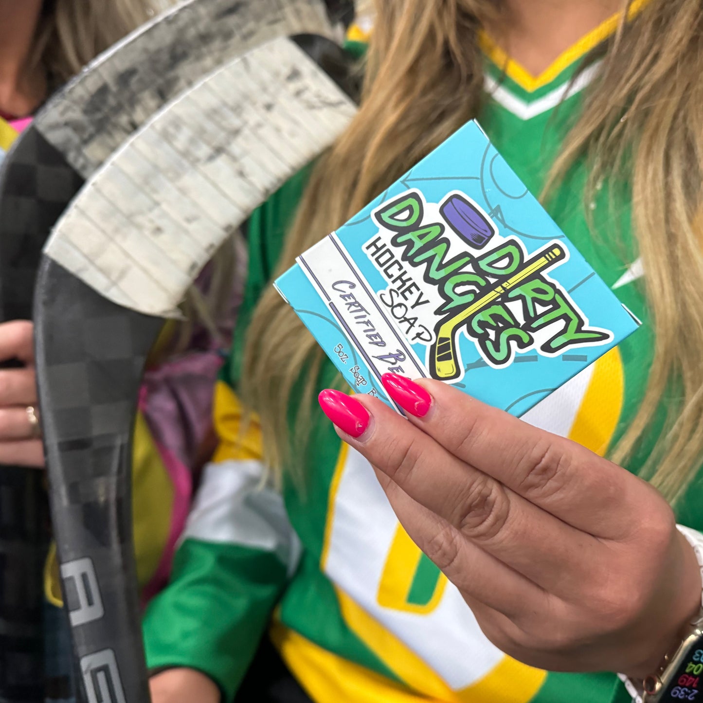 a woman hockey player holding a blue box of dirty dangles hockey soap certified beaut scent inside an ice rink.
