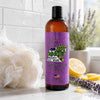 Bottle of Dirty Dandies body wash with a purple label on a bathroom counter, surrounded by lemons and lavender.