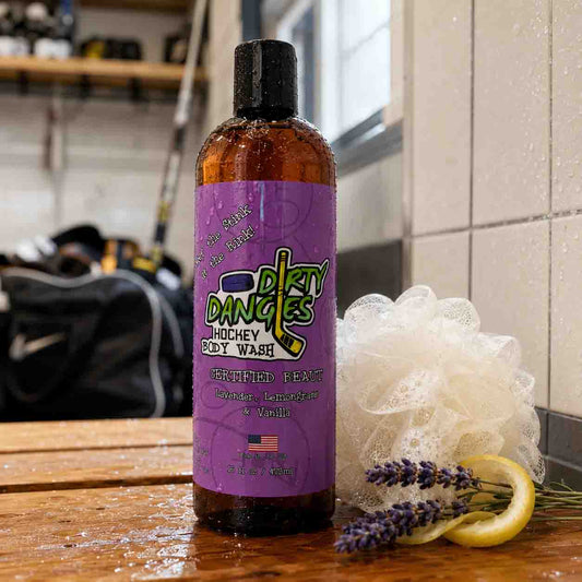 Bottle of Dirty Dangles hockey body wash on a wooden surface with a soapy sponge and lavender flowers.