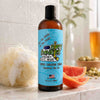 Bottle of Dirty Dangles body wash on a bathroom counter with a loofah and grapefruit.