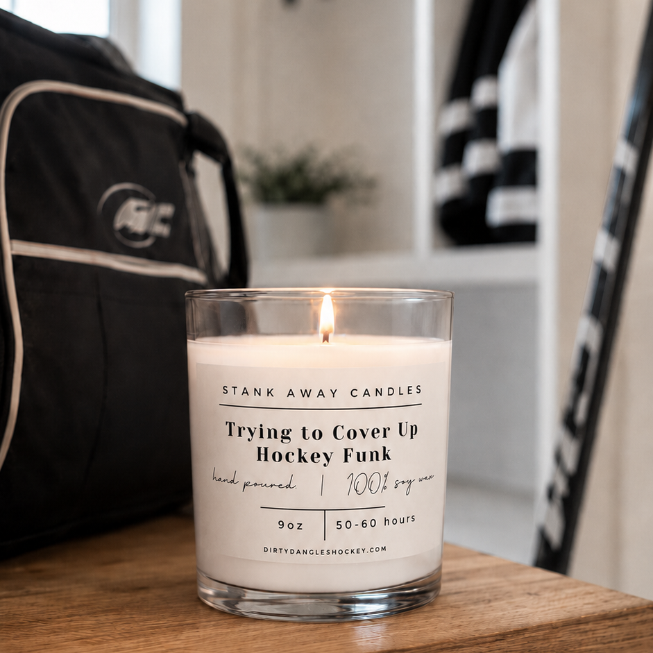 A white scented hockey candle on a wood bench with hockey gear. Trying to cover up hockey funk