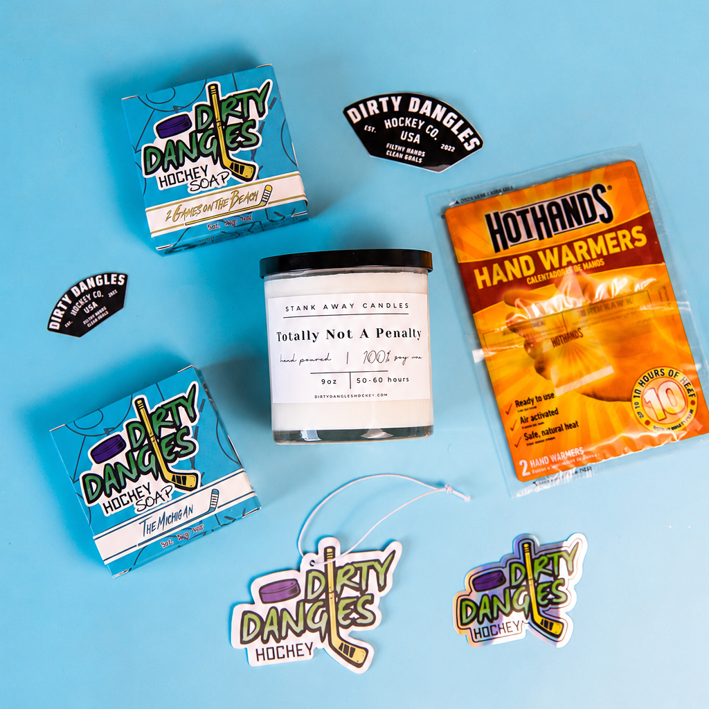 A styled flat-lay of multiple products on a light blue background, including two blue boxes of Dirty Dances Holistic 'Healing Day' supplements, a white jar candle labeled 'Honestly Not A P*sshole', an orange packet of HotHands Hand Warmers, and several die-cut stickers.