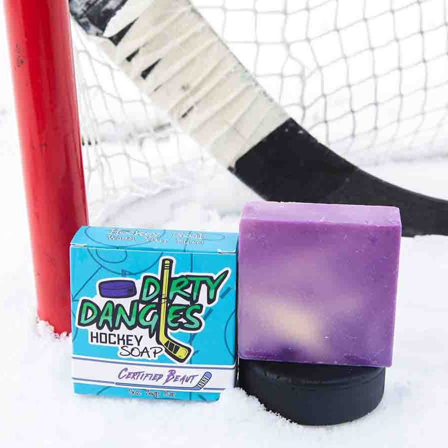 A purple and white soap bar sits in the snow with a hockey goal, hockey puck and a hockey stick. Certified Beaut