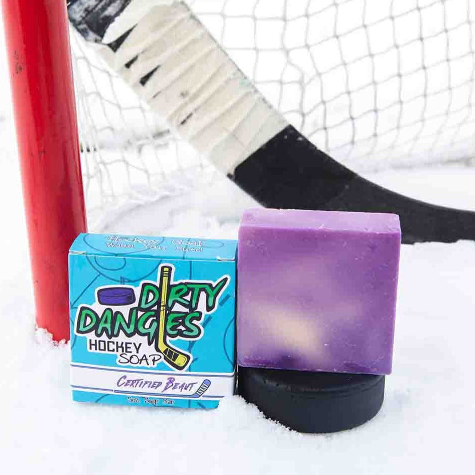 A purple and yellow soap bar sits in the snow with a hockey goal, hockey puck and a hockey stick. Certified Beaut scent