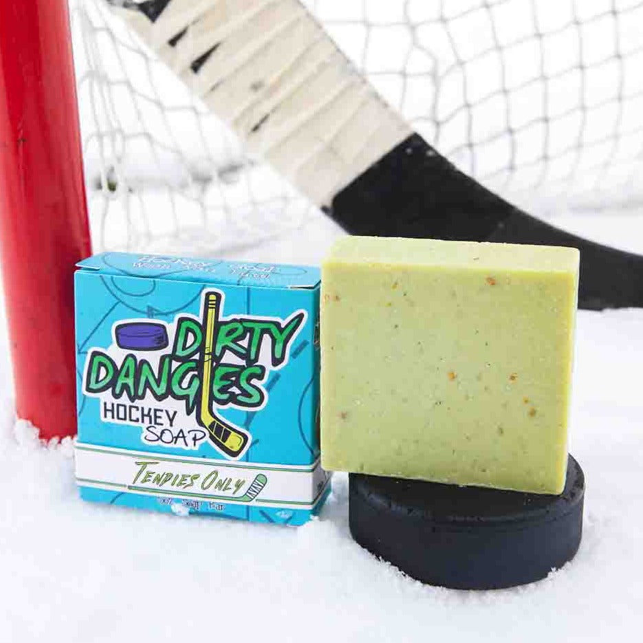 A green soap bar sits in the snow with a hockey goal, hockey puck and a hockey stick. Tendies Only scent