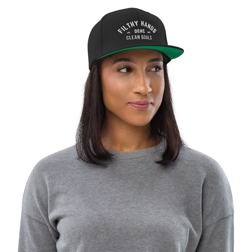 A woman in a black flat brim hat with a green underbrim on white background.. Filthy hands clean goals