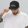 A man wearing a A black flat brim hat on a gray background. Dirty dangles hockey co.