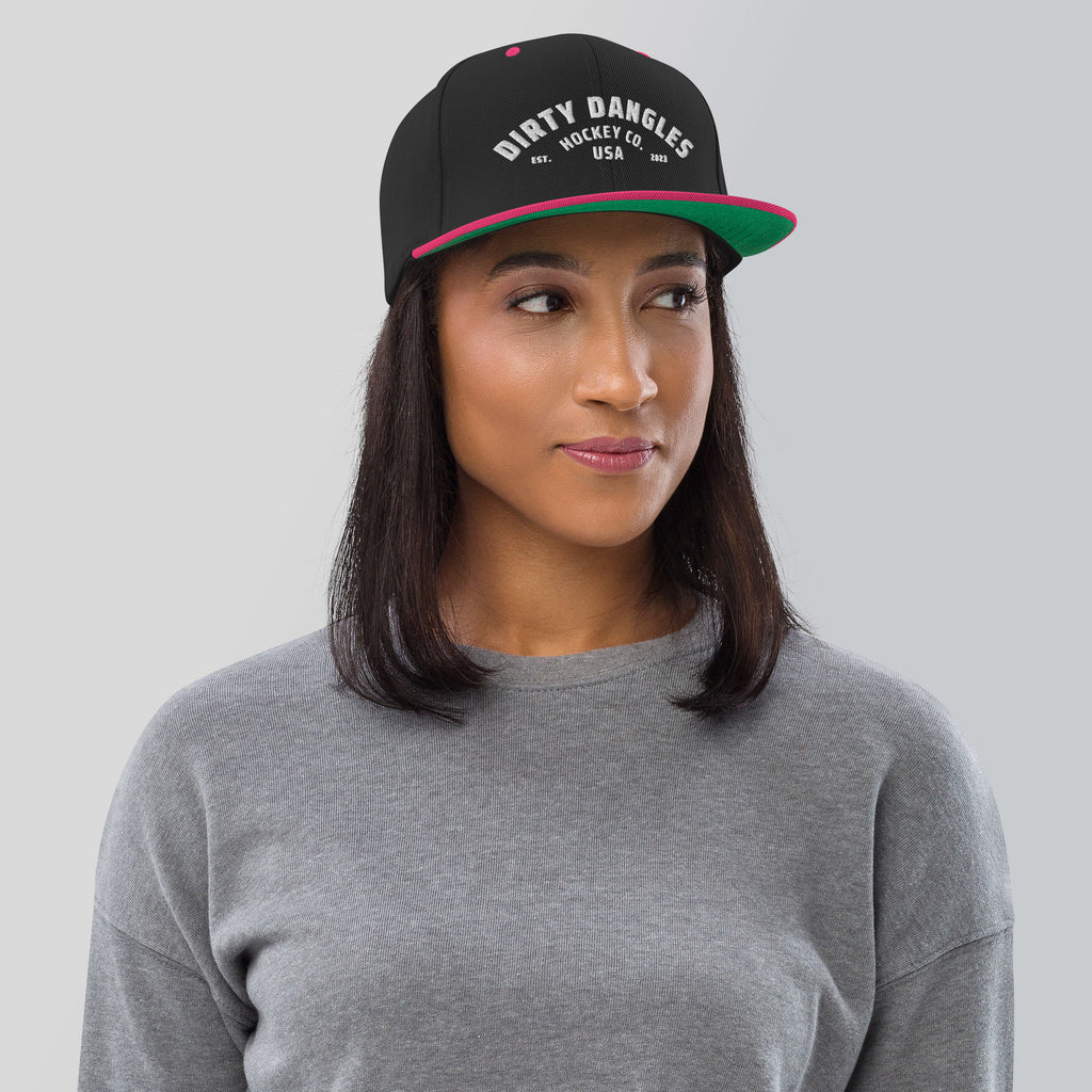 A woman wearing  a black and pink flat brim hat on a gray background. Dirty dangles hockey co.