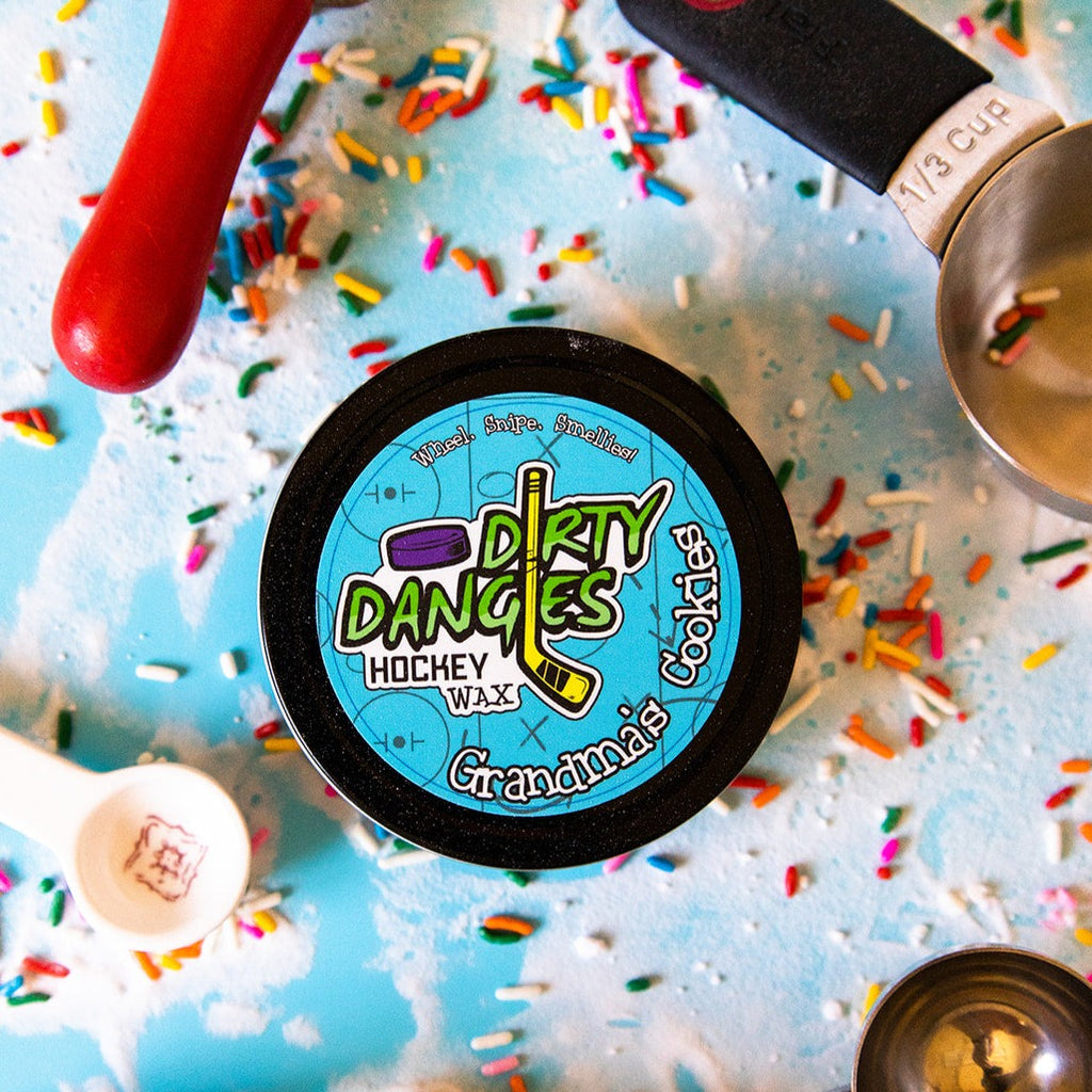 A tin of Dirty Dangles Hockey Stick Wax Grandma's Cookies Scent sits on a blue background surrounded by sprinkles and baking supplies