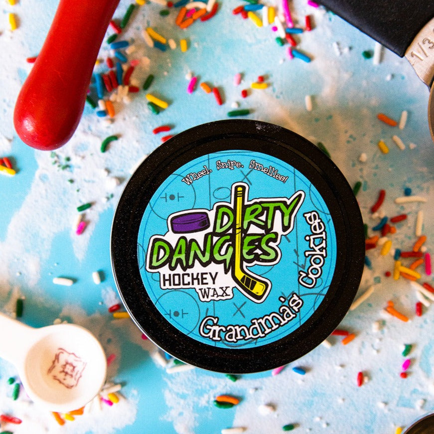 A tin of Dirty Dangles Hockey Stick Wax Grandma's Cookies Scent sits on a blue background surrounded by sprinkles and baking supplies