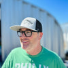 A man outside smiling wearing a light gray and black dirty dangles hockey co. mesh snapback trucker hat.