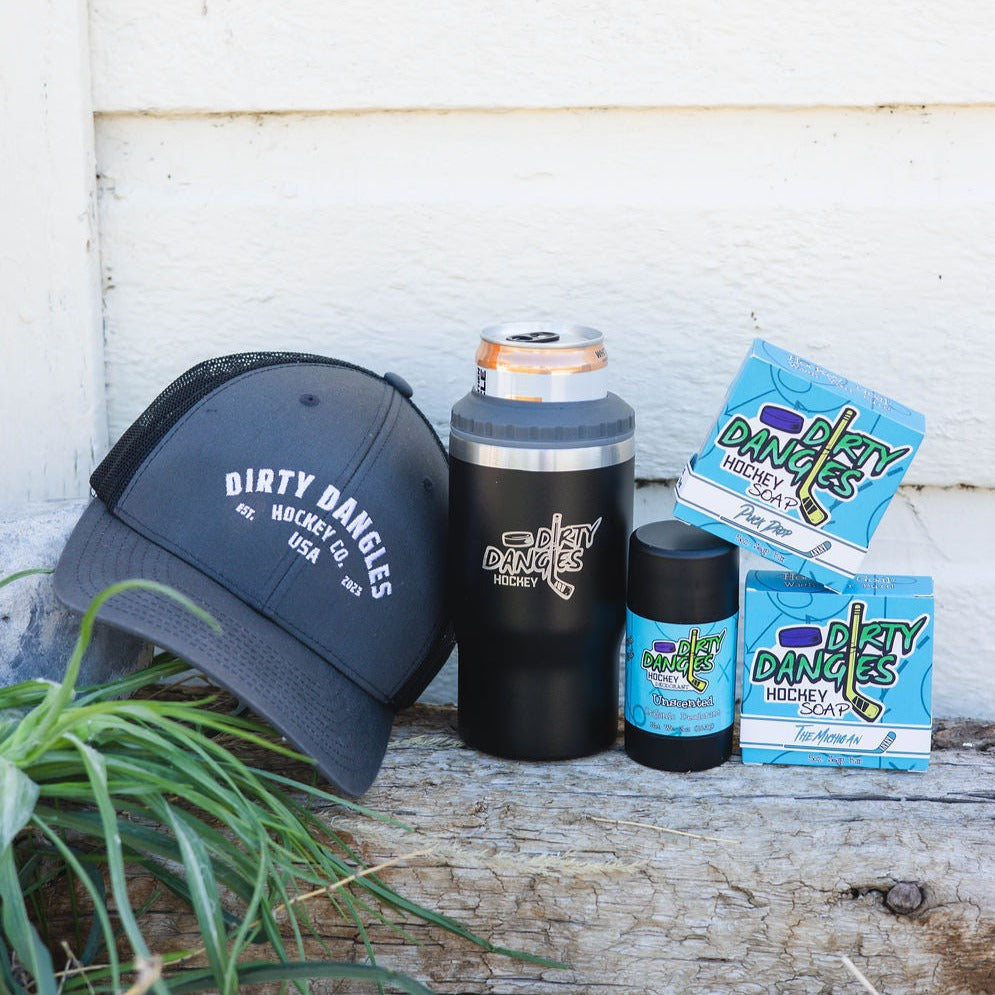 2 bars of hockey soap, a black 2 in 1 drink tumbler can holder, a stick of dirty dangles natural deodorant and a dirty dangles hockey co. hat outside on a piece of wood.
