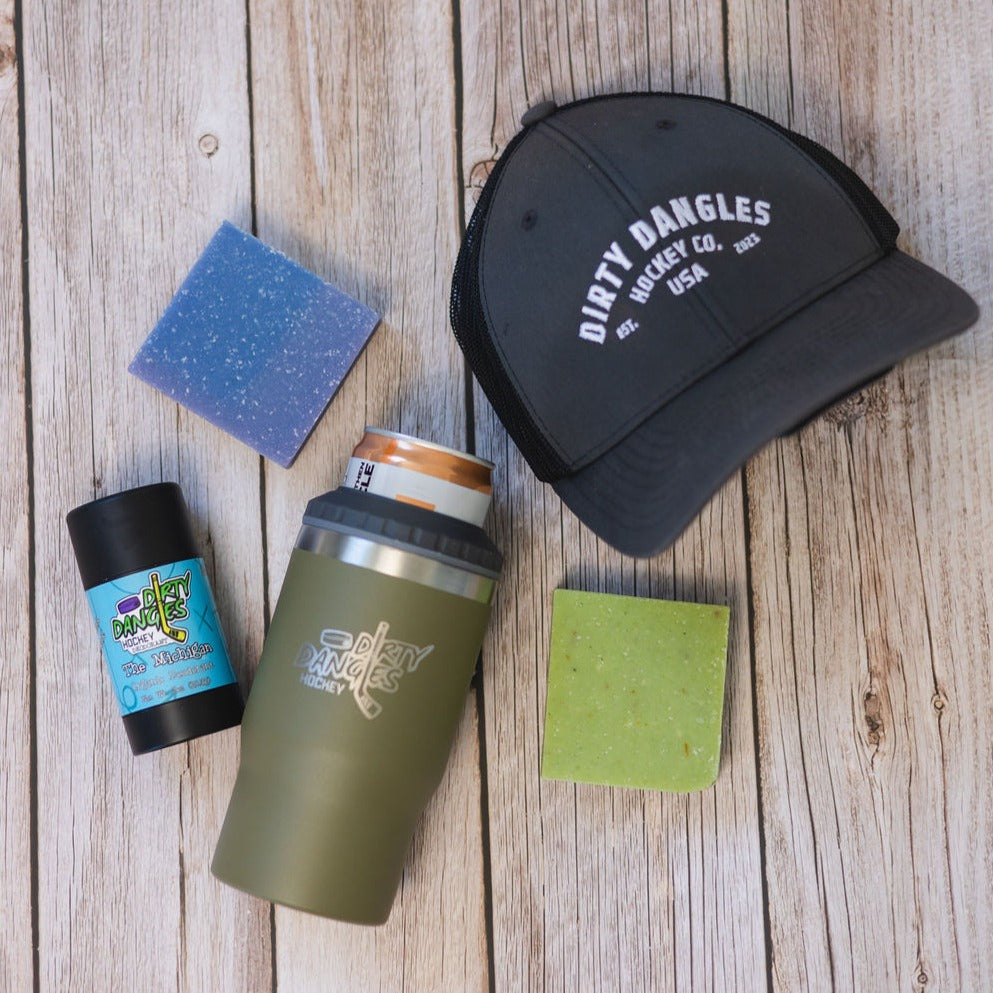 2 bars of hockey soap, a Tactical Green 2 in 1 drink tumbler can holder, a stick of dirty dangles natural deodorant and a dirty dangles hockey co. hat on a wood background.