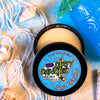 A tin of Dirty Dangles Hockey Stick Wax Lava Monster Scent sits open on a blue background surrounded by sand, seashells and a tiki mug