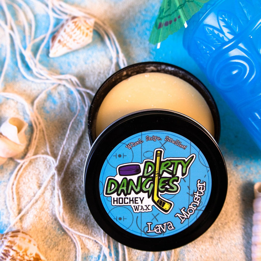 A tin of Dirty Dangles Hockey Stick Wax Lava Monster Scent sits open on a blue background surrounded by sand, seashells and a tiki mug