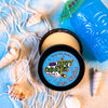 A tin of Dirty Dangles Hockey Stick Wax Lava Monster Scent sits open on a blue background surrounded by sand, seashells and a tiki mug