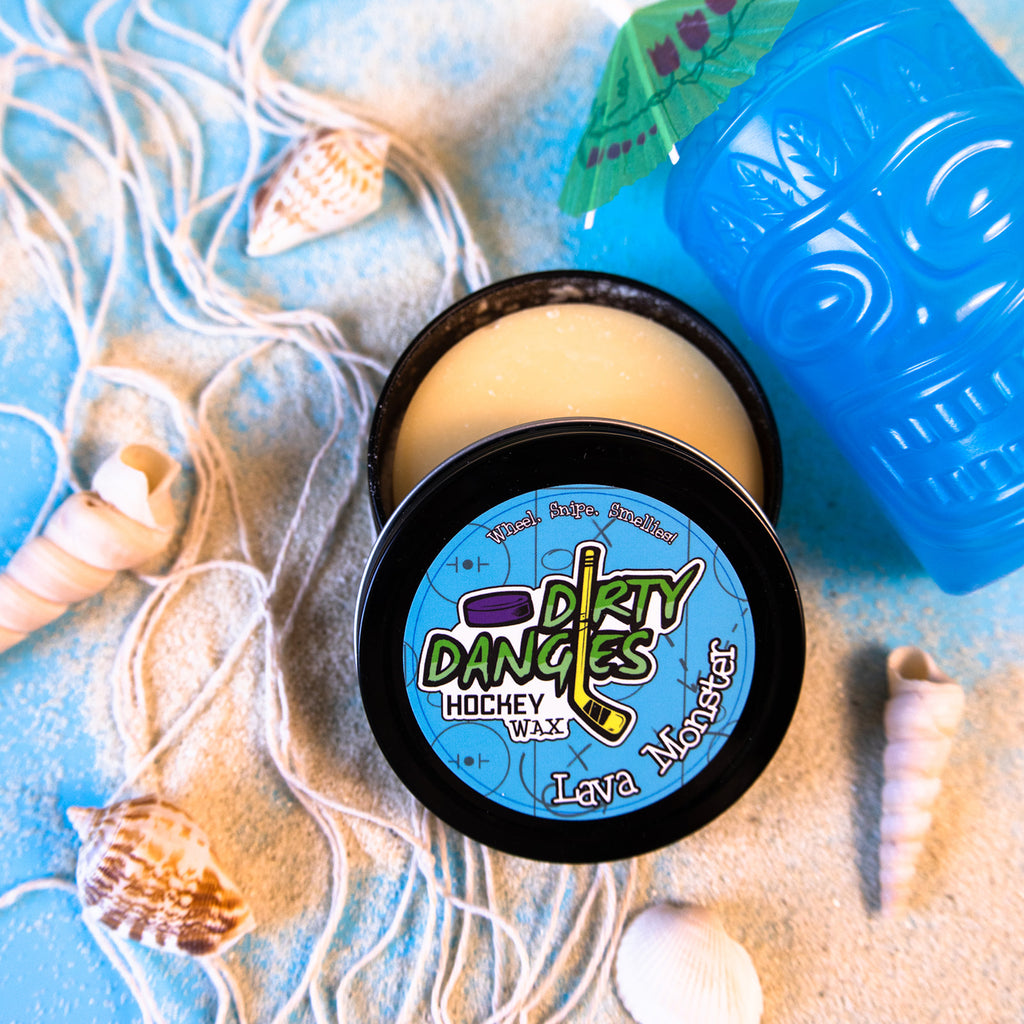 A tin of Dirty Dangles Hockey Stick Wax Lava Monster Scent sits open on a blue background surrounded by sand, seashells and a tiki mug