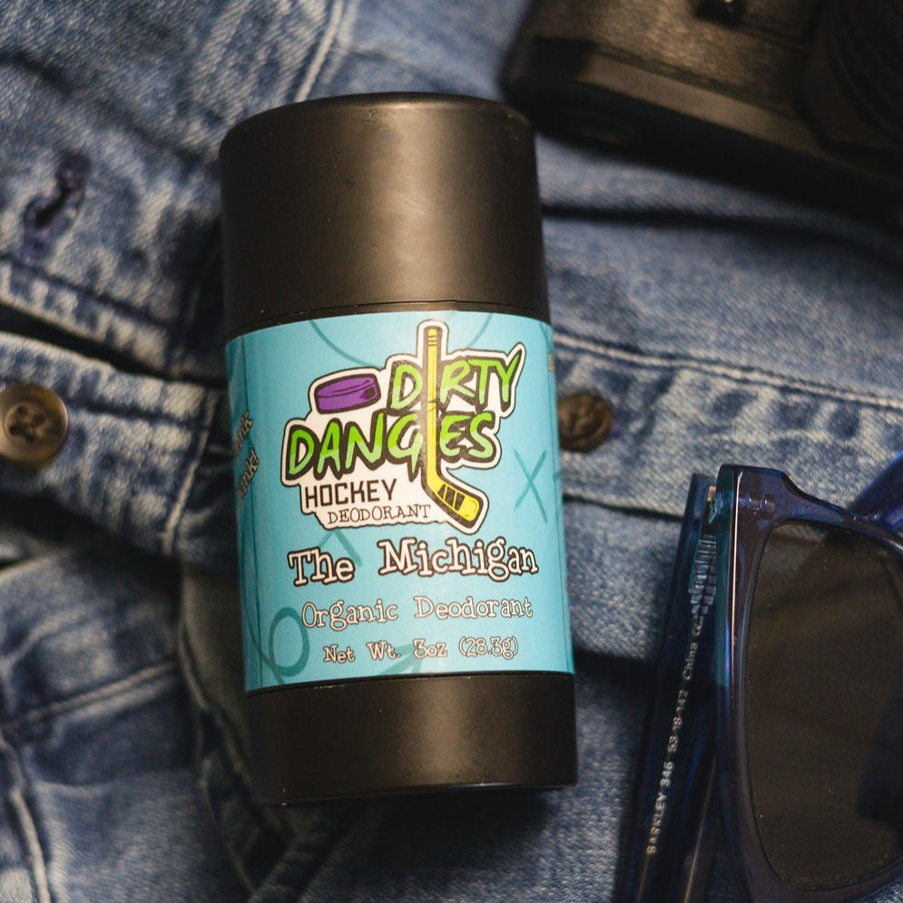 A tube of dirty dangles natural probiotic deodorant on a blue background with jeans, sunglasses and a camera