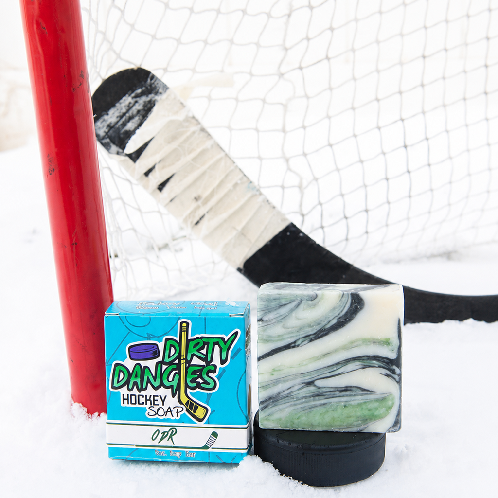 A handmade soap bar with a green and white marble pattern sits on a black hockey puck next to its blue and white 'Dirty Dangles Hockey Soap' box, set in the snow in front of a hockey goal and stick.