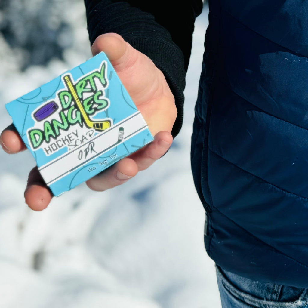 A hockey player holding a box of Dirty Dangles ODR soap near an outdoor ice rink. A green, black and white swirl soap bar,