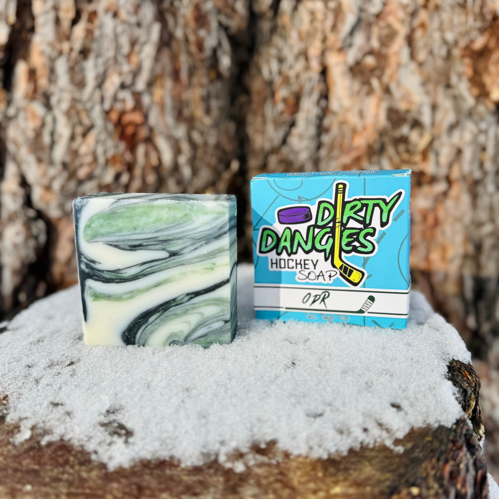 A bar and box of Dirty Dangles ODR soap in front of a snowy tree trunk. A green, black and white swirl soap bar,