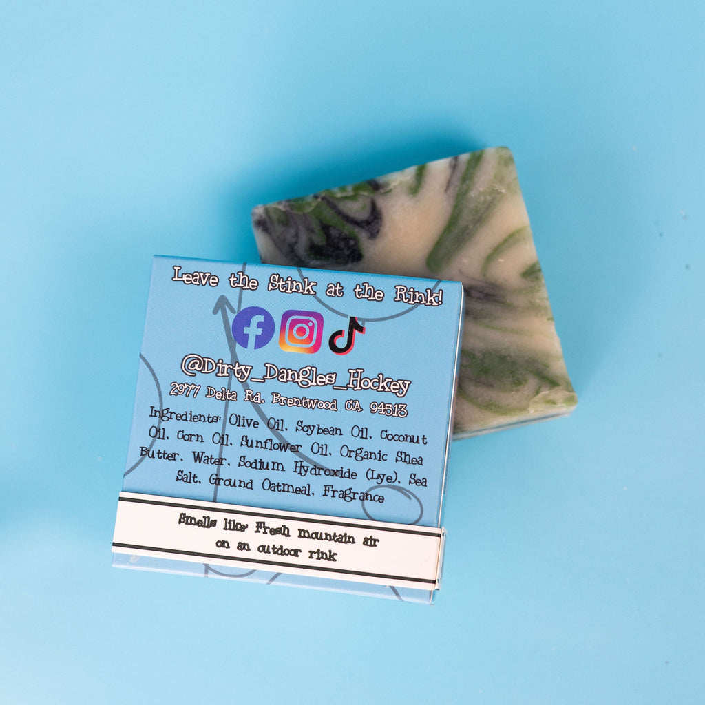 A bar and back of a box of Dirty Dangles ODR soap on the boards of an outdoor ice rink. A green, black and white swirl soap bar. Smells like fresh mountain air on an outdoor rink.