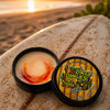 An open tin of dirty dangles tiki deke limited edition tropical stick wax on a rustic surfboard at the beach