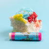 A pink stick of dirty dangles natural lip balm. Vanilla scent on a blue background with a piece of vanilla cake.