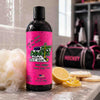 A bottle of Dirty Dangles Girl's Hockey Body Wash in goal digger lemon sugar cookie scent on a locker room bench with cookies, lemon slices and a white loofah with a girl's hockey bag in the background.
