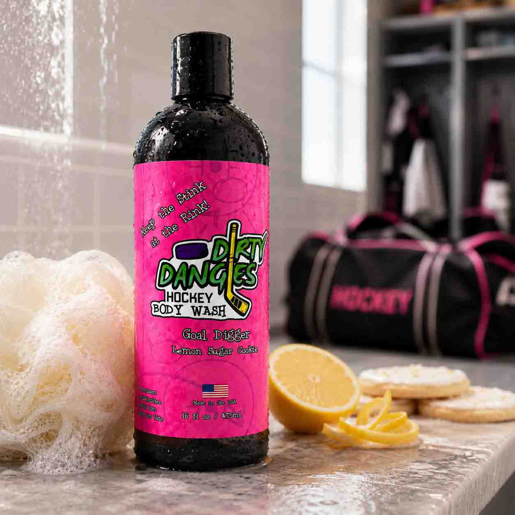A bottle of Dirty Dangles Girl's Hockey Body Wash in goal digger lemon sugar cookie scent on a locker room bench with cookies, lemon slices and a white loofah with a girl's hockey bag in the background.