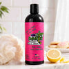 A bottle of Dirty Dangles girl's Hockey Body Wash goal digger lemon sugar cookie scent with a bright pink label and black cap, standing on a bathroom counter with a loofah, lemon slices, and sugar cookies in a bright bathroom setting.