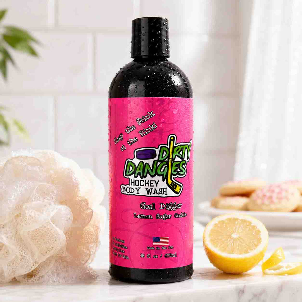 A bottle of Dirty Dangles girl's Hockey Body Wash goal digger lemon sugar cookie scent with a bright pink label and black cap, standing on a bathroom counter with a loofah, lemon slices, and sugar cookies in a bright bathroom setting.