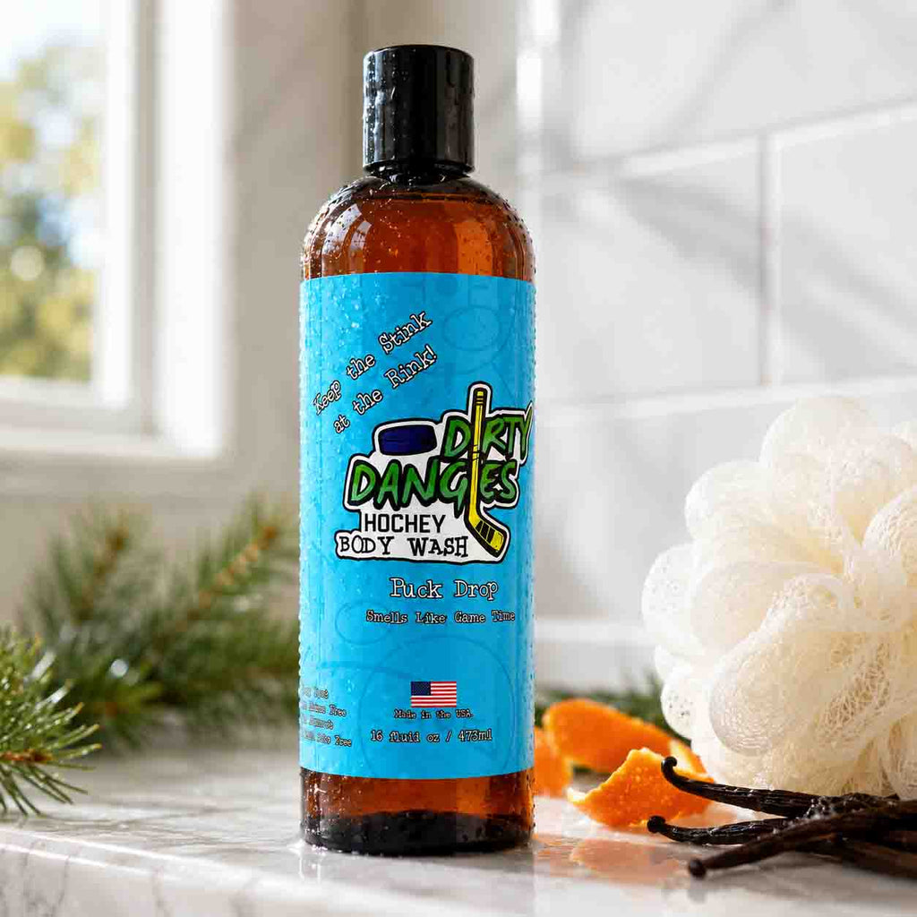 A bottle of Dirty Dangles Hockey Body Wash in puck drop scent, featuring a bright blue label and black cap, set on a marble countertop with a loofah, orange peel, pine needles and vanilla beans in a bathroom setting.