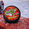 Container of Dirty Dangles waffle sauce scent hockey wax on a snowy surface with a hockey helmet in the background.