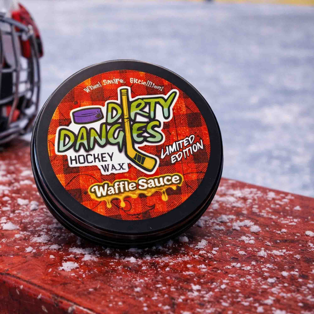 Container of Dirty Dangles waffle sauce scent hockey wax on a snowy surface with a hockey helmet in the background.