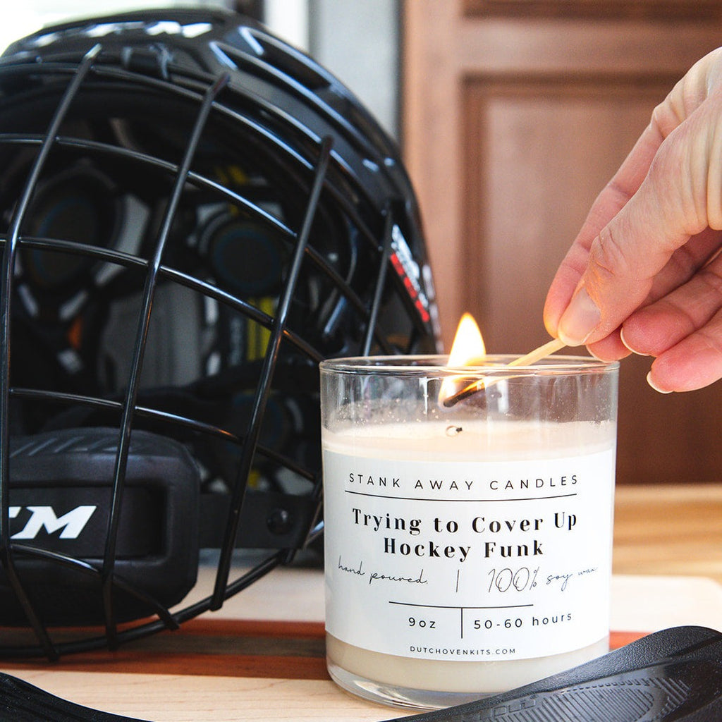 A white candle  sits on a table with a hockey stick and a hockey helmet. A hand lights the candle with a match. Trying to cover up hockey funk