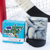 A black and white swirl soap bar sits in the snow with a hockey goal, hockey puck and a hockey stick. Sneaky Dekes