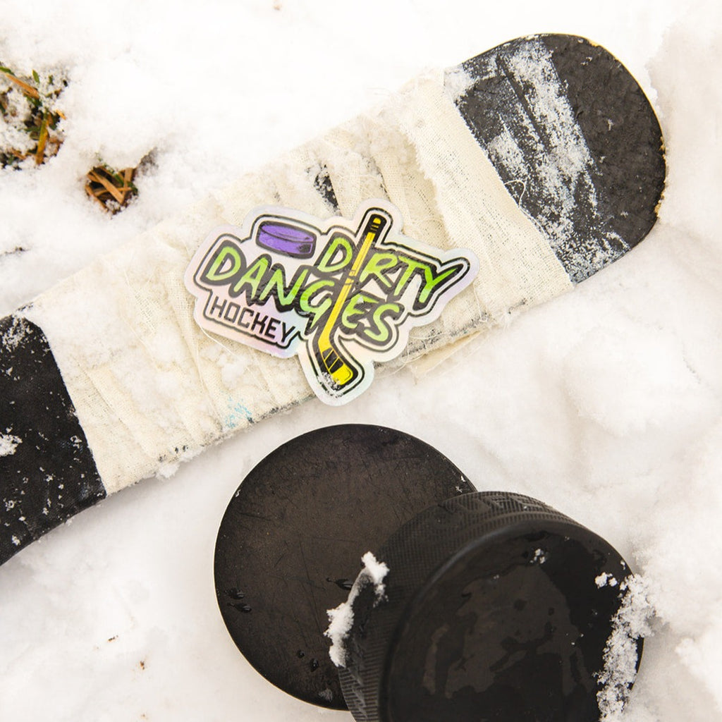 A dirty dangles hockey logo sticker sits on a hockey stick in the snow with 2 hockey pucks. Dirty dangles hockey.