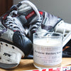 A white candle sits on a table with a  pair of ice skates and a hockey stick. Because Hockey Gear Natural Soy Wax Candle