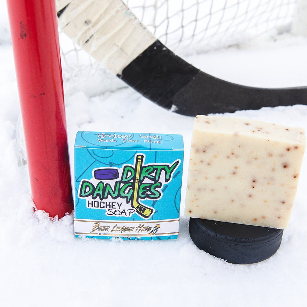 A white and orange beer league hero grapefruit pale ale soap bar sits in the snow with a hockey goal, hockey puck and a hockey stick.