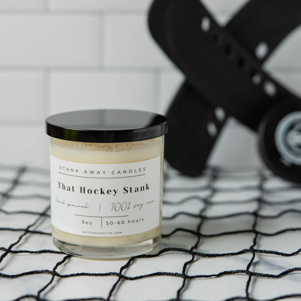 A white candle sits on a black hockey net with hockey sticks and a hockey puck. That hockey stank
